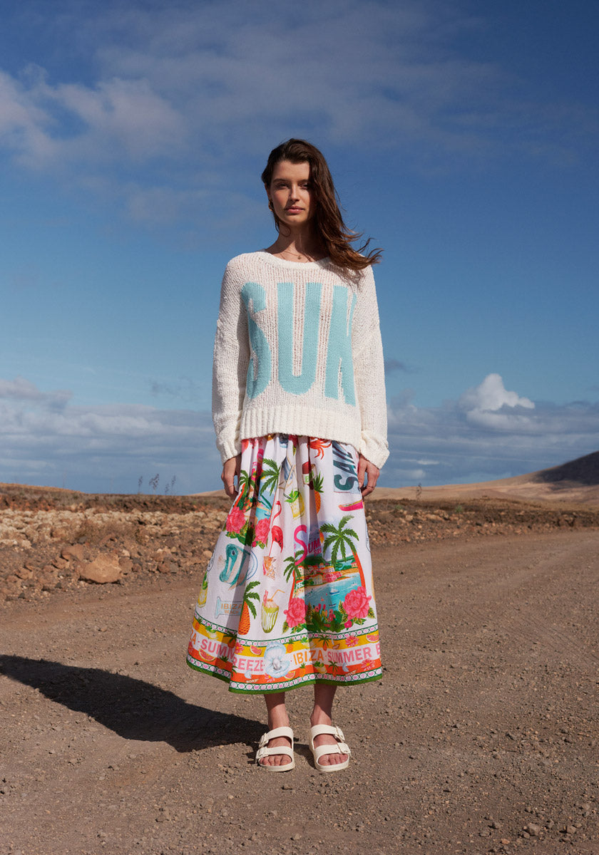 A woman stands on a dirt road under a partly cloudy sky, wearing the Frogbox by Princess Pullover Sun with large aqua lettering and a colorful tropical skirt. She also wears white sandals and looks directly at the camera.