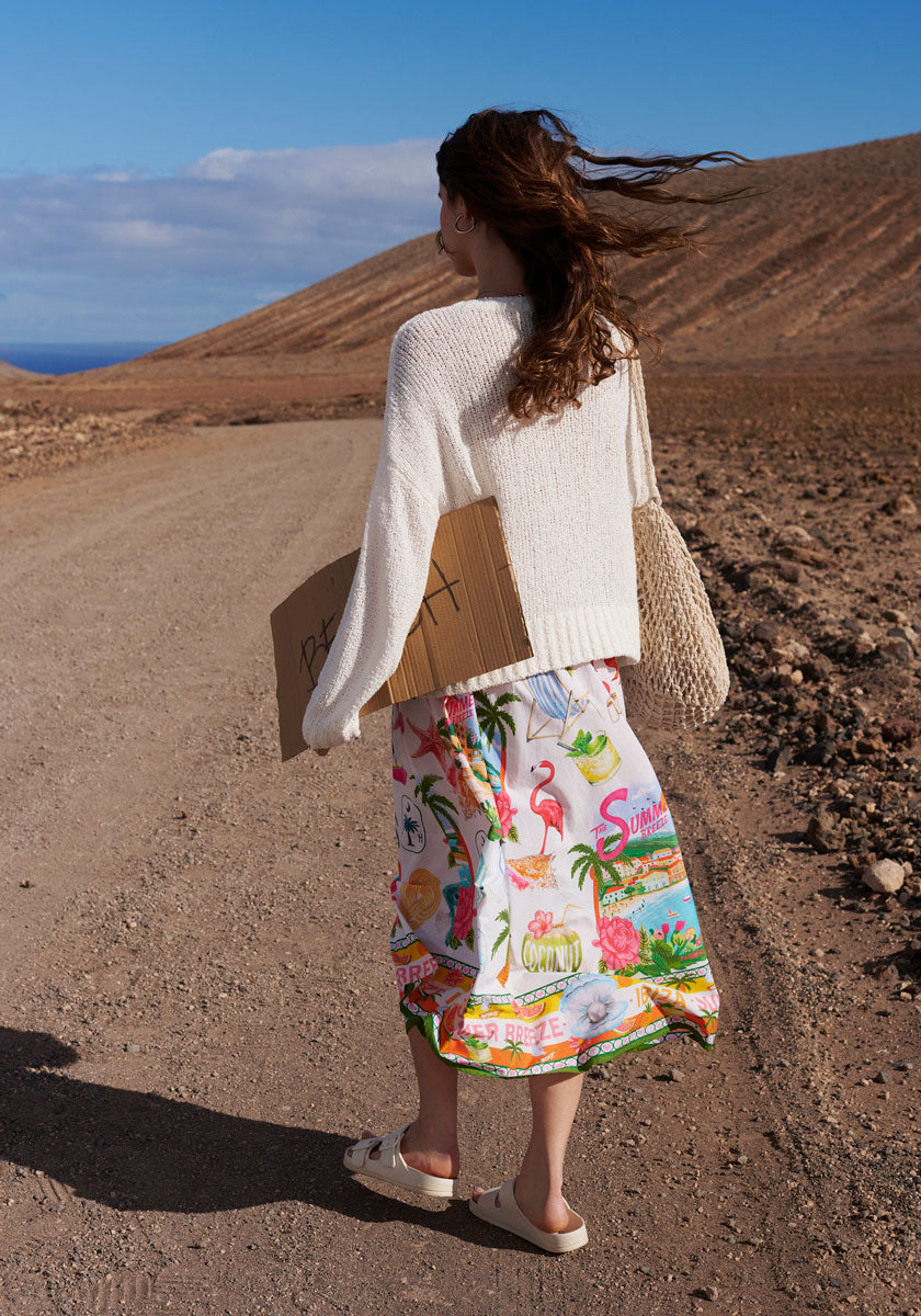 A woman with long brown hair, wearing the Frogbox by Princess Pullover Sun and a colorful tropical skirt, stands on a dirt road holding a crochet bag and cardboard sign, gazing toward distant hills under the blue sky.
