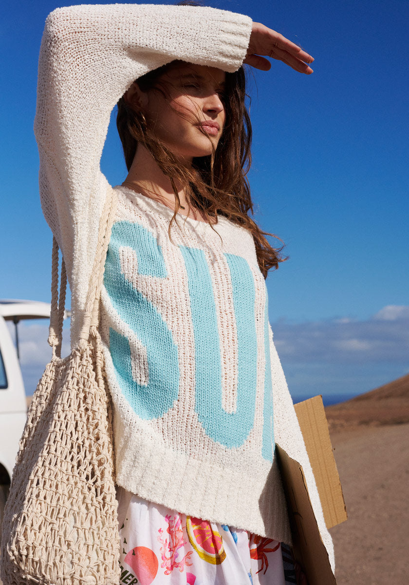 A woman stands outdoors on a sunny day, shielding her eyes. She wears the Frogbox by Princess Pullover Sun aus Baumwolle-Kaschmir-Mischung mit großem türkisen Sun-Schriftzug, eine Netz-Tasche und einen bunten Rock. Ein Van und Hügel sind im Hintergrund.