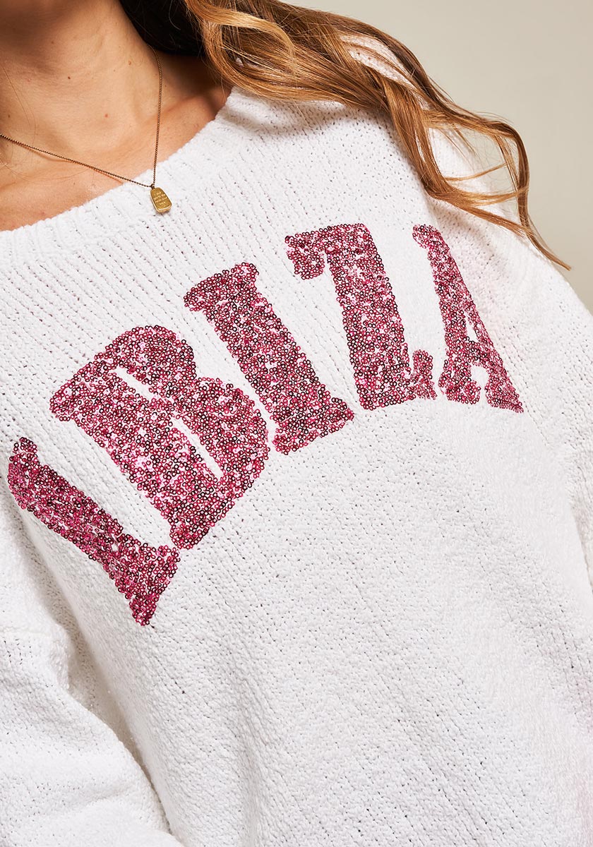 A woman in a textured white Frogbox by Princess organic cotton pullover with IBIZA in large pink sequins on the chest. Her wavy brown hair, gold necklace, and shoulder are visible against a plain, neutral background.
