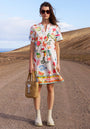A woman stands on a rocky desert road, wearing the Frogbox by Princess Blusenkleid Summer Breeze with a summer print, cream ankle boots, sunglasses, and a woven bag. The dress hem reads “IBIZA SUMMER BREEZE.” She gazes neutrally at the camera.