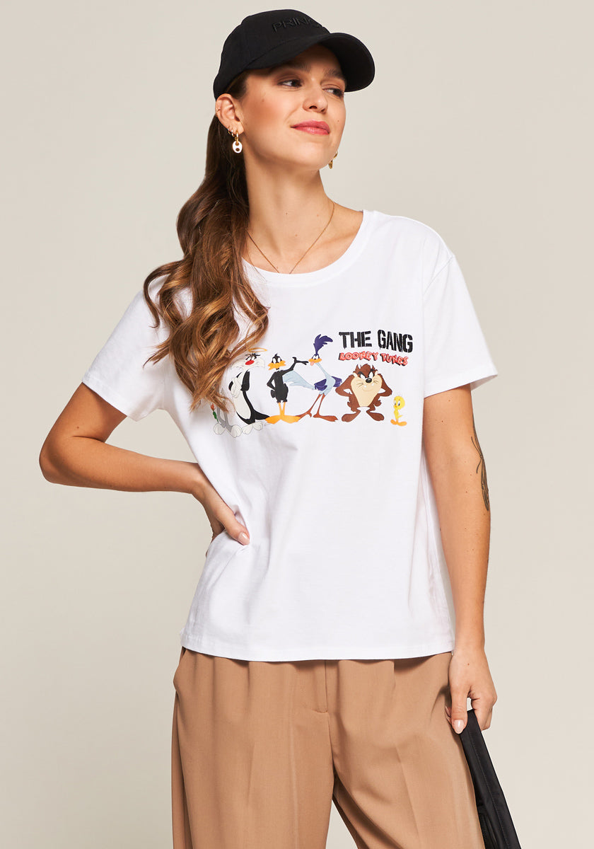 A woman with wavy brown hair wears a black cap, gold hoop earrings, a white T-shirt featuring Looney Tunes characters, and beige pants. She stands confidently with one hand on her hip and a slight smile, against a plain light background.
