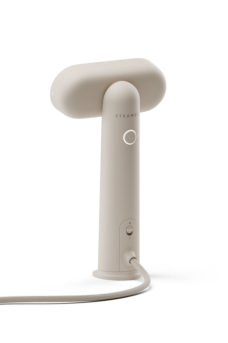 Ein beiges tragbares Dampfglätter von Steamery mit einem T-förmigen Oberteil, einem runden Einschaltknopf, einer kleinen Kontrollleuchte und einer stoffbezogenen Kordel an der Basis. Dieser minimalistische Kleidungsdampfer steht aufrecht auf einem weißen Hintergrund mit einem modernen Design.