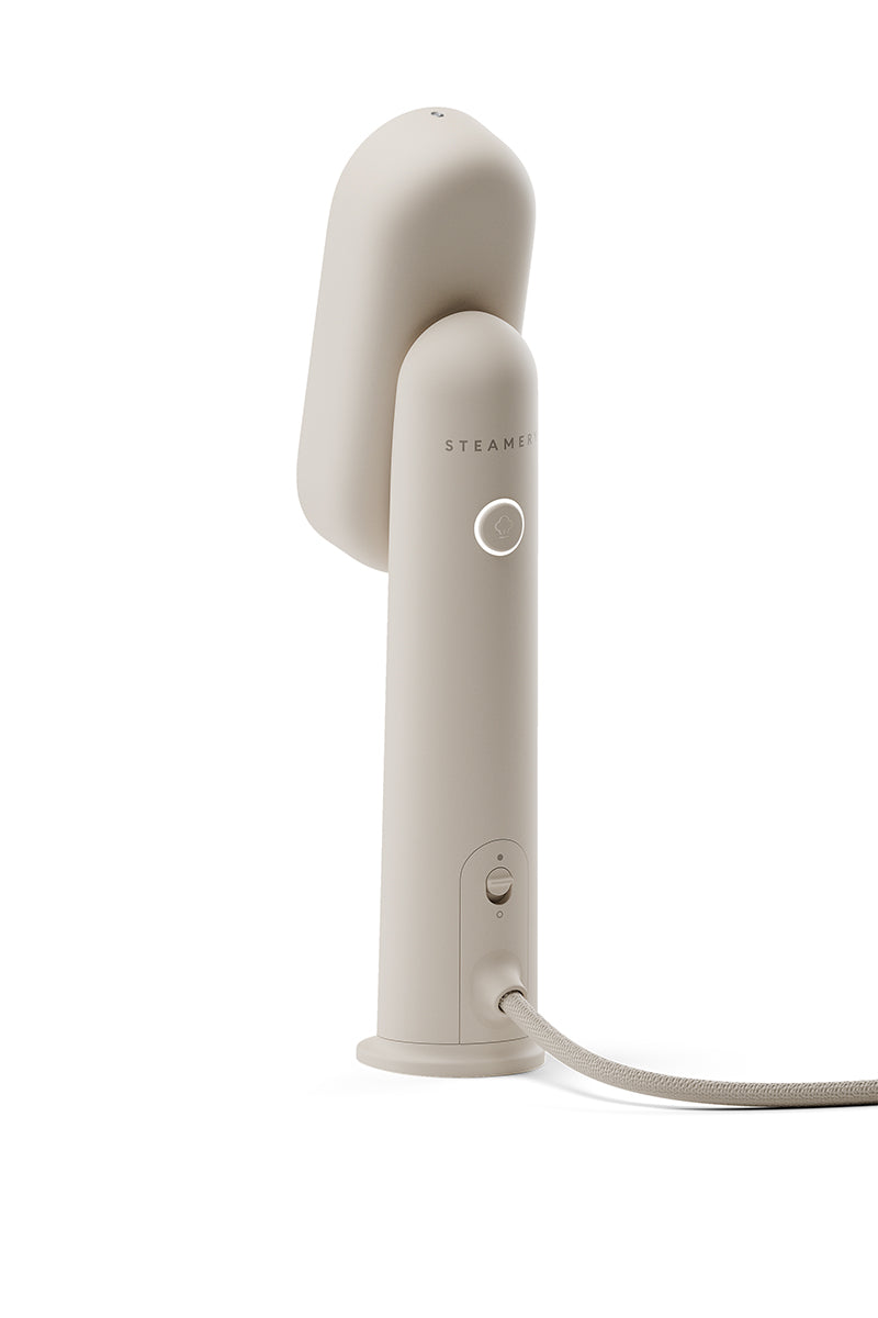 Ein beiger tragbarer Dampfbügler von Steamery mit modernem, minimalistischem Design steht aufrecht auf weißem Hintergrund. Er hat eine glatte, abgerundete Oberseite, einen mit STEAMERY beschrifteten Knopf und eine am Boden befestigte Stoffkordel.