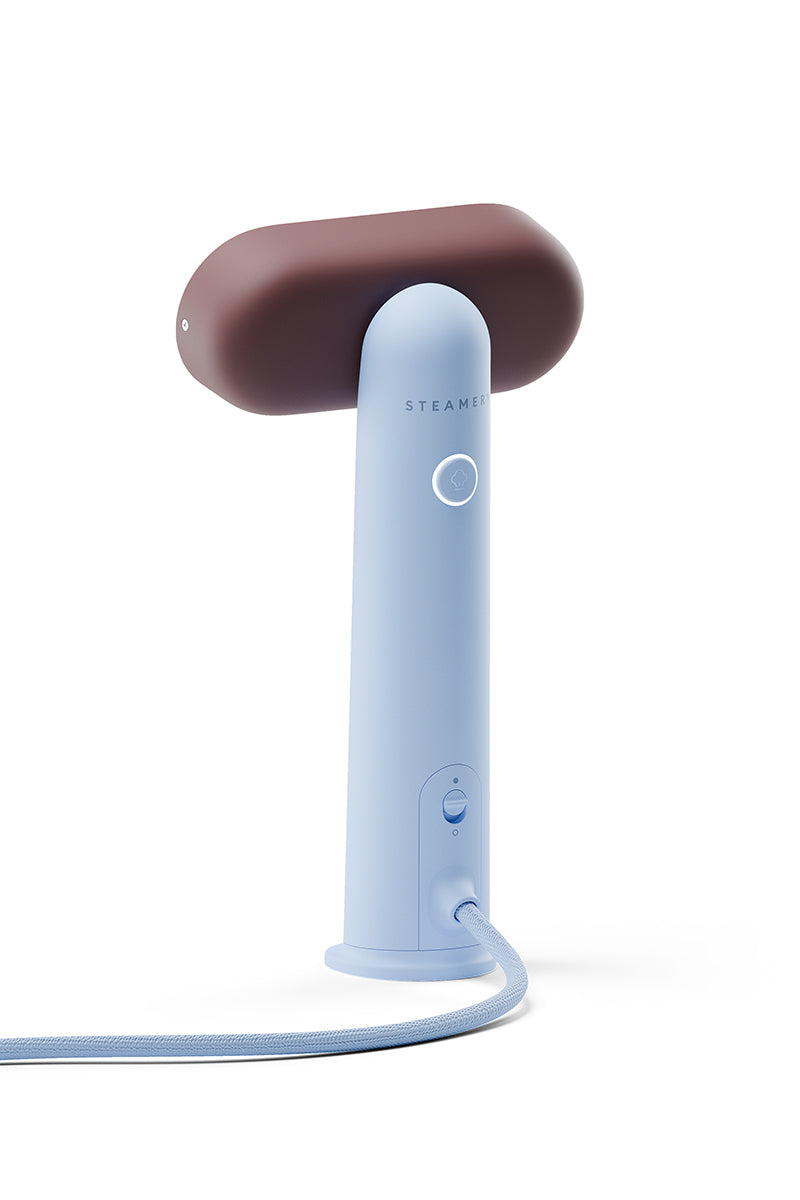 Der Steamery tragbarer Dampfglätter hat ein schlankes, hellblaues, zylindrisches Design mit einem abgerundeten braunen Deckel. Dieser Reisesteamer steht aufrecht auf einem flachen Sockel, hat ein unteres Netzkabel und einen runden Einschaltknopf, der in der Mitte leuchtet.