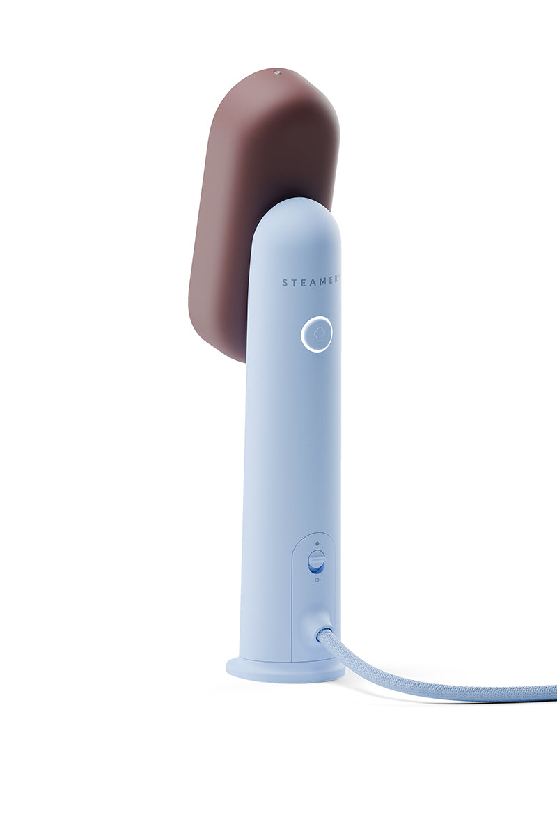 Der Steamery Reisesteamer ist ein moderner, minimalistischer tragbarer Dampfbügler mit einem hellblauen Gehäuse, einem braunen Dampfkopf, einem leuchtenden, kreisrunden Einschaltknopf und einer Stoffkordel, alles vor einem strahlend weißen Hintergrund.