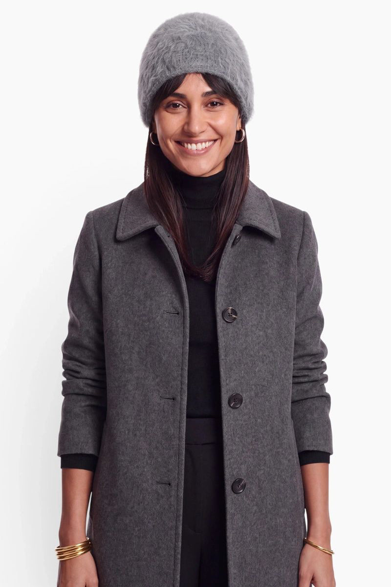Eine lächelnde Frau mit langen dunklen Haaren trägt eine flauschige graue Kaschmir-Mütze, einen schwarzen Rollkragenpullover und einen dunkelgrauen Wollmantel. Als Accessoires trägt sie goldene Creolen und goldene Armreifen an beiden Handgelenken.