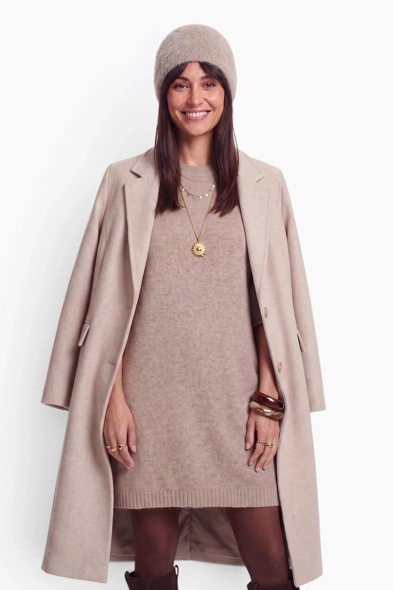 Eine lächelnde Frau trägt ein beiges Strickkleid, einen dazu passenden Mantel, der ihr über die Schultern drapiert ist, und eine flauschige Kaschmir-Mütze. Als Accessoires trägt sie mehrreihige Goldketten mit einem Sonnenanhänger sowie dunkle Armbänder vor einem einfarbigen, hellcremefarbenen Hintergrund.