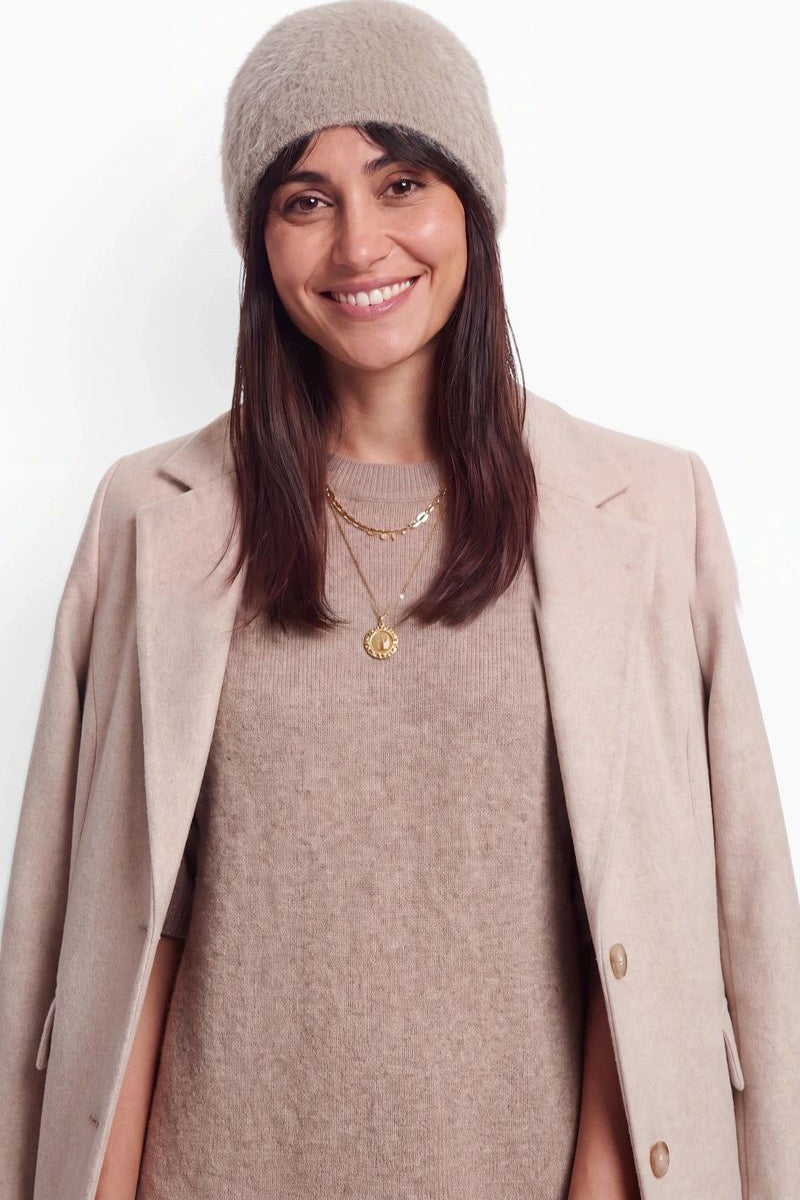 Eine lächelnde Frau mit langen braunen Haaren trägt eine flauschige beige Kaschmir-Mütze, einen dazu passenden Pullover und einen hellrosa Mantel. Vor einem schlichten weißen Hintergrund trägt sie zwei übereinanderliegende goldene Halsketten.