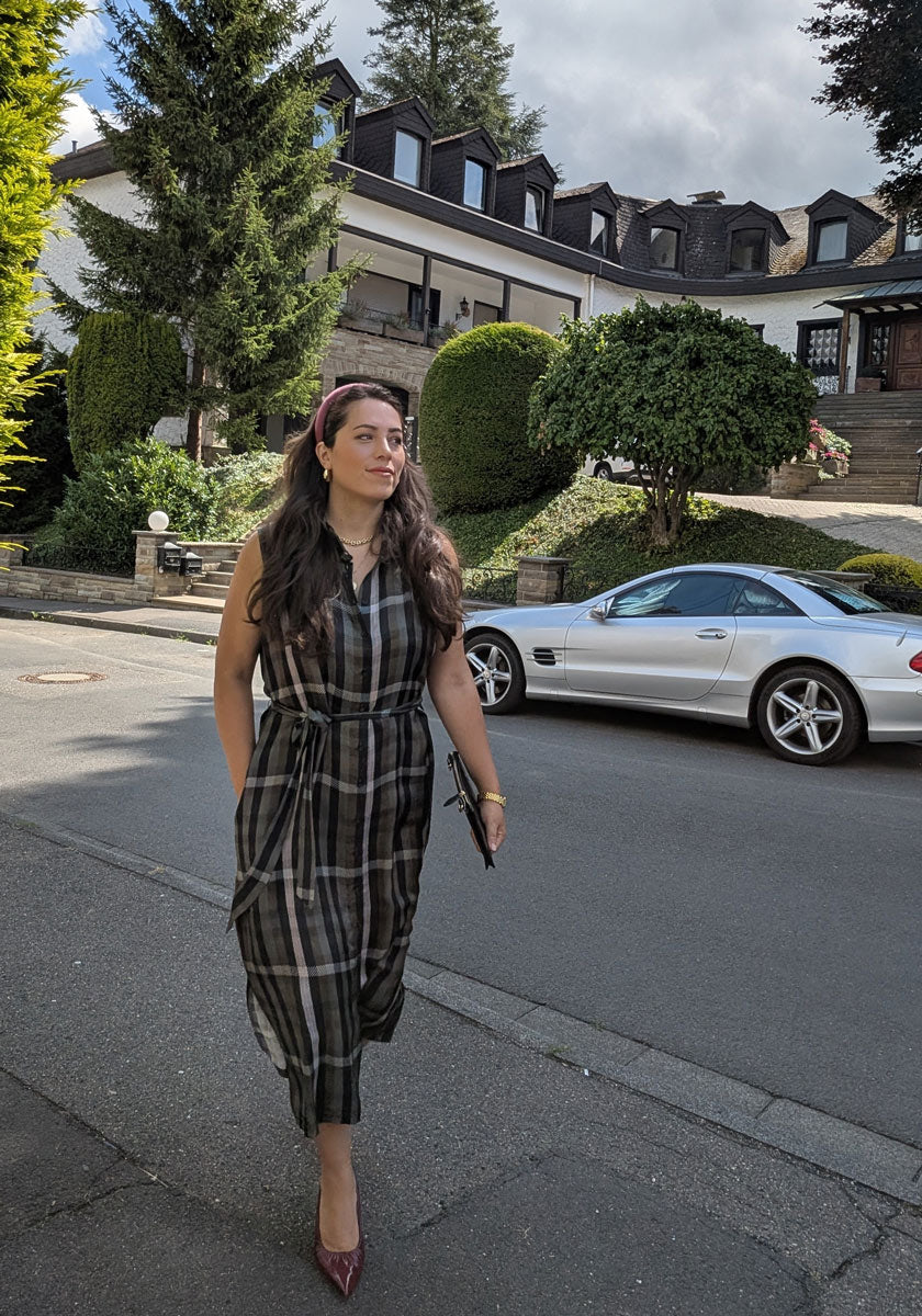 A woman with long dark hair in a Princess Goes Hollywood plaid Midikleid with Seideanteil and red shoes walks along a green-lined street, clutch in hand and serious expression. A white convertible sports car is parked before a large house behind her.