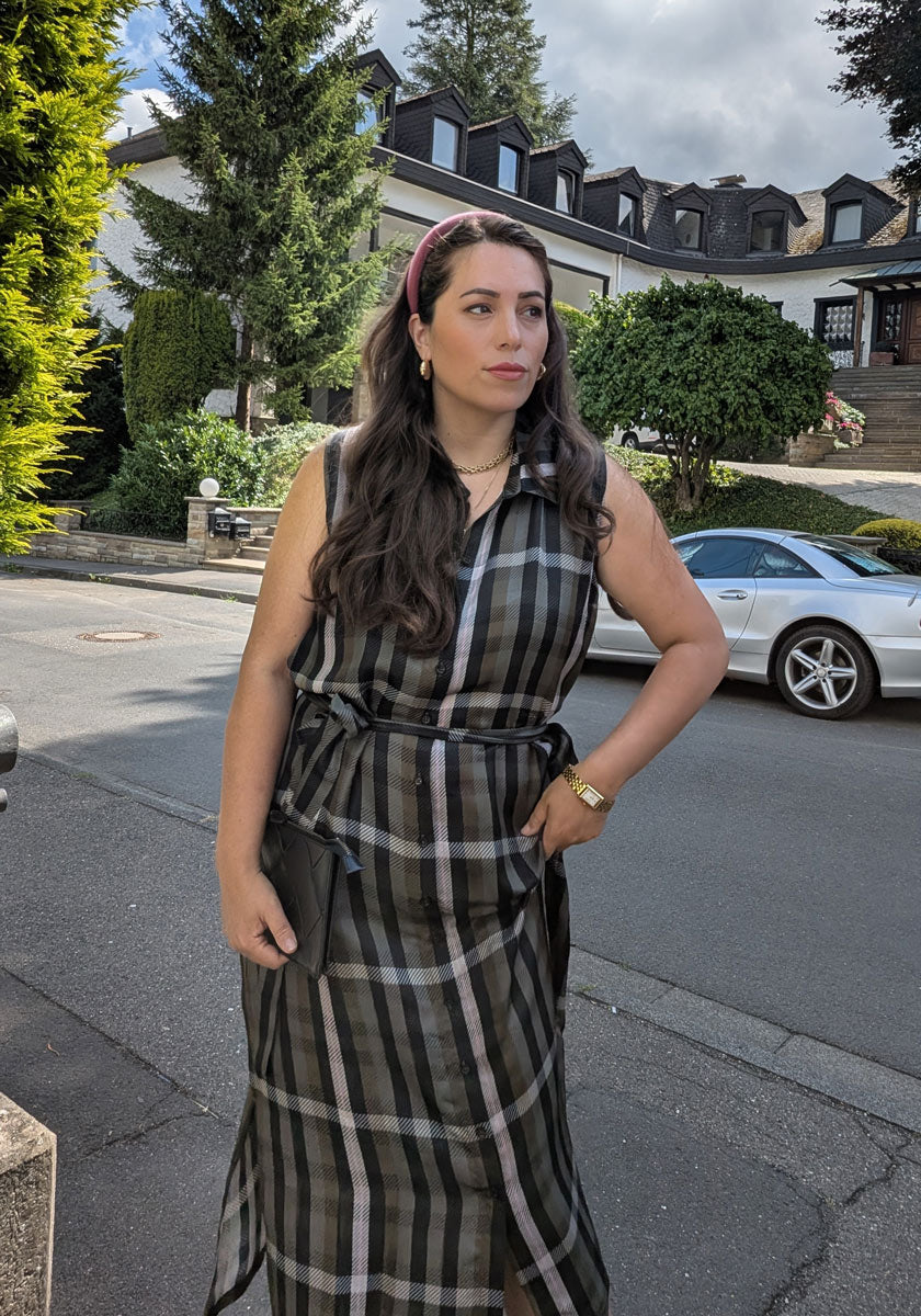 A woman with long wavy brown hair, in a pink headband and a sleeveless Princess Goes Hollywood black check-print midi dress with tie belt, stands on a residential street, holding a black clutch. A white sports car and large house are behind her.