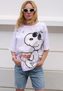 A woman with blond hair wears white cat-eye sunglasses, a loose Princess Goes Hollywood Snoopy T-shirt with a large Snoopy graphic holding a rainbow guitar, light denim shorts, and a pink watch as she stands against a white wall.