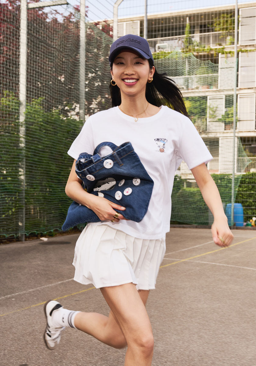 A young woman runs on an outdoor court, smiling in a Princess Goes Hollywood look: navy cap, white T-shirt, pleated skirt, sneakers, and a Denim-Shopper with pins. Trees and an apartment building are in the background.