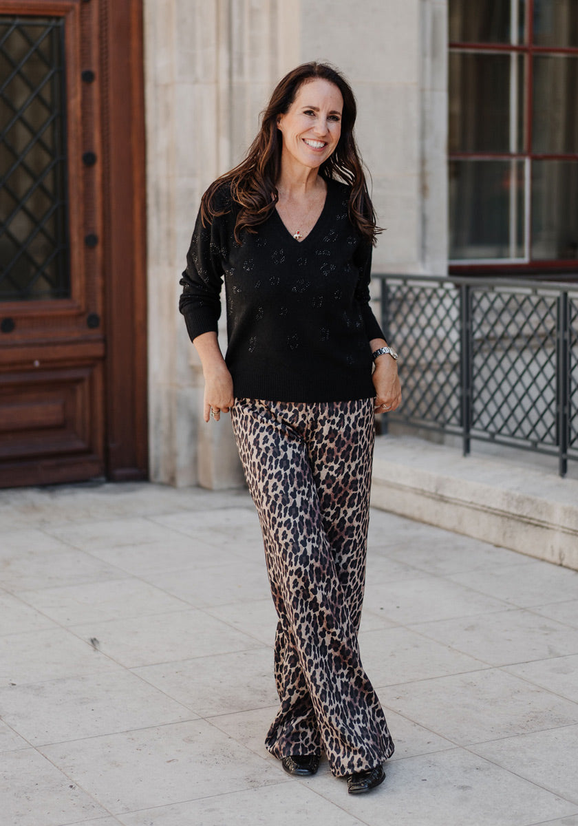 A woman with long brown hair smiles outdoors, wearing a Princess Goes Hollywood black V-Ausschnitt Strickpullover with subtle patterns and loose leopard-print pants, standing on light stone pavement near a large wooden door and metal railing.
