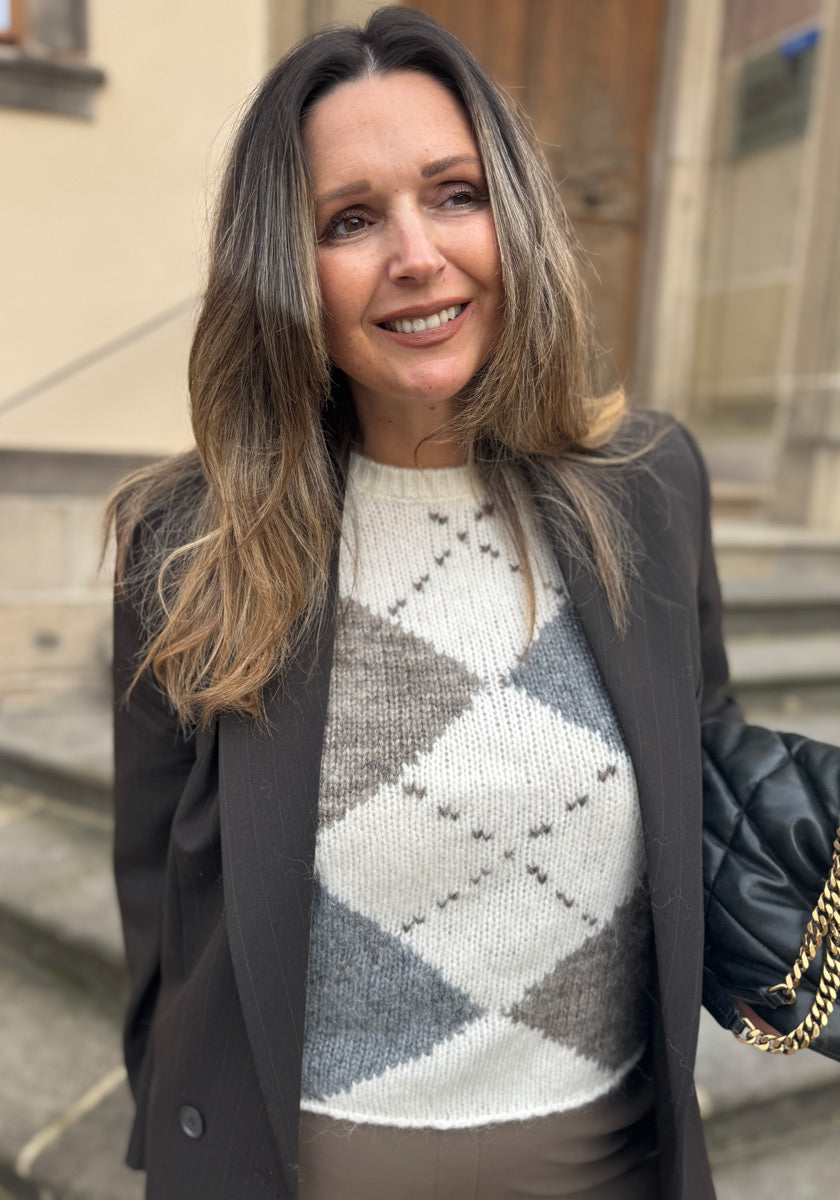 Eine Frau mit langen, gewellten braunen Haaren steht draußen auf den Steinstufen und lächelt. Sie trägt einen dunklen Blazer über einem Princess Goes Hollywood Woll-Alpaka Argyle Strickpullover und hat eine schwarze gesteppte Handtasche mit einer Goldkette in der Hand.