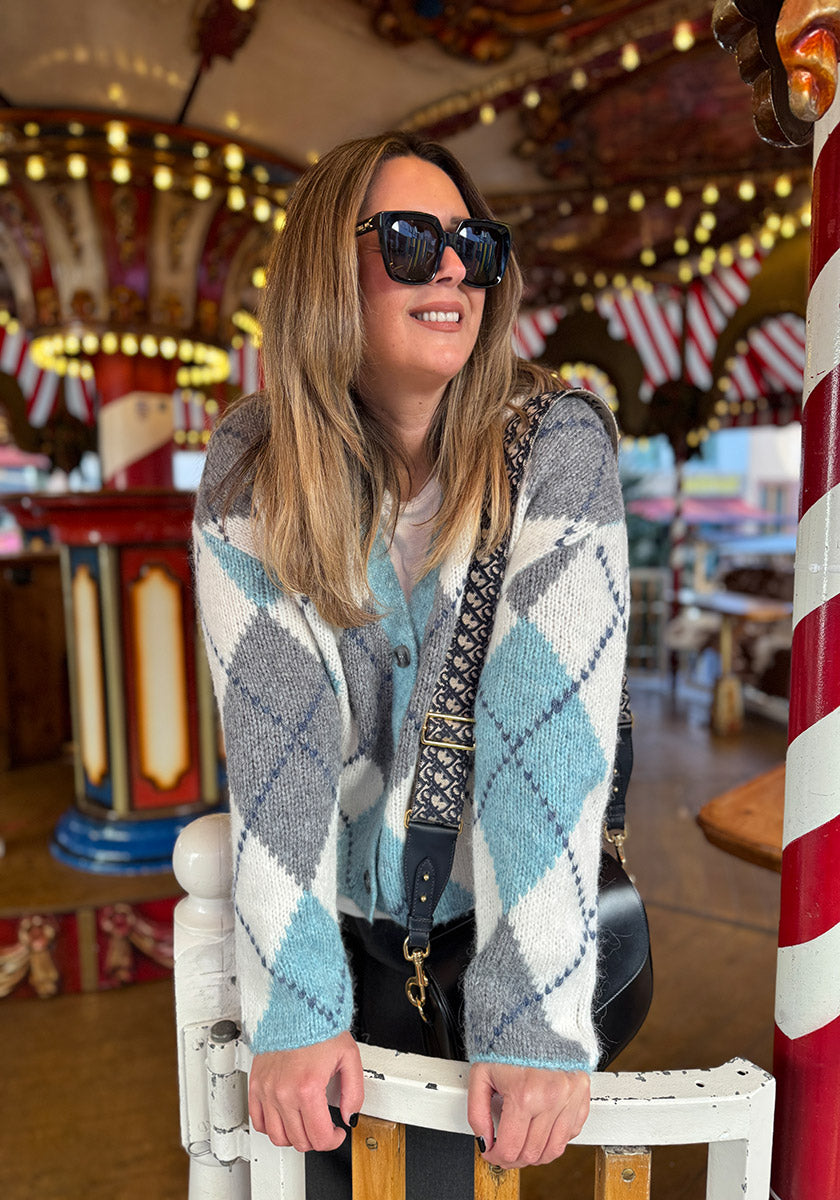 Eine Frau mit langen blonden Haaren und Sonnenbrille lächelt, während sie sich an einen weißen Zaun lehnt. Sie trägt eine blau-weiß-graue Argyle-Muster Strickjacke aus Wolle-Alpaka-Mix von Princess Goes Hollywood vor einem bunten rot-weißen Karussell.