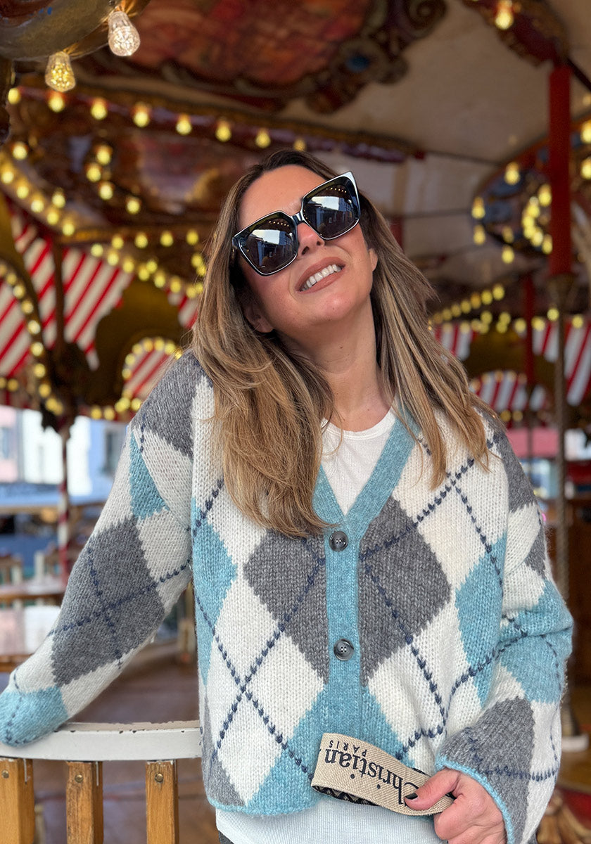 Eine Frau mit übergroßer schwarzer Sonnenbrille und einer blau-grau-weißen Princess Goes Hollywood"-Woll-Alpaka-Strickjacke lächelt vor einem hell erleuchteten Karussell mit roten und weißen Streifen und hält einen kleinen Gegenstand mit der Aufschrift Christian".