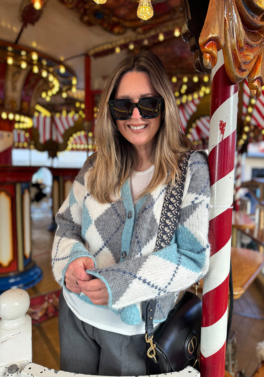 Eine Frau mit langem, gesträhntem Haar und schwarzer Sonnenbrille lächelt vor einem Karussell. Sie trägt eine Princess Goes Hollywood Argyle-Muster Strickjacke aus Wolle-Alpaka-Mix über einem weißen Oberteil und trägt eine schwarze Tasche mit gemustertem Riemen.