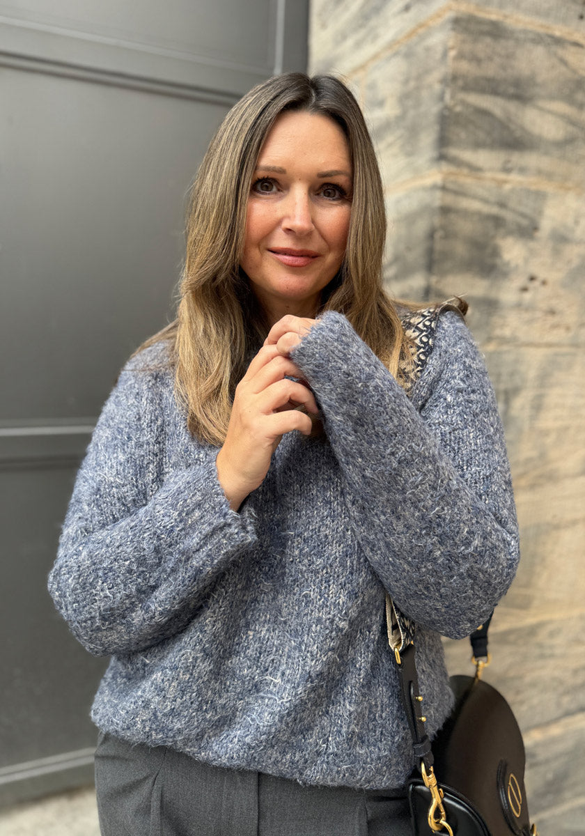 Eine Frau mit langen, gesträhnten Haaren trägt einen graublauen flauschigen Pullover von Princess Goes Hollywood und eine graue Hose. Sie steht an einer Steinmauer, hält eine schwarze Handtasche mit einem goldenen Kettenriemen und lächelt sanft.