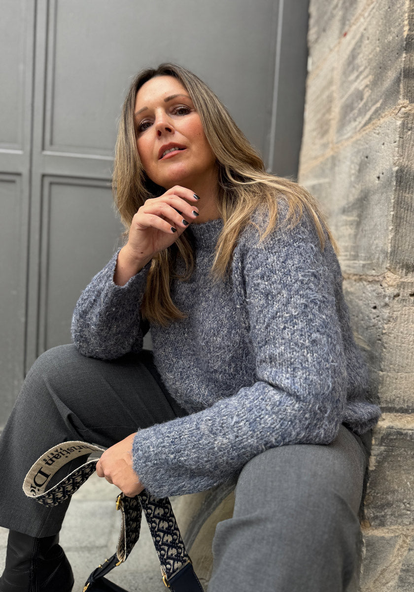 Eine Frau mit langen blonden Haaren sitzt auf Stufen an einer grauen Steinmauer. Sie trägt eine graue Hose und einen Princess Goes Hollywood Luxus Strickpullover. Sie hält eine marineblau gemusterte Tasche in der Hand und blickt selbstbewusst in die Kamera, eine Hand neben ihrem Gesicht.