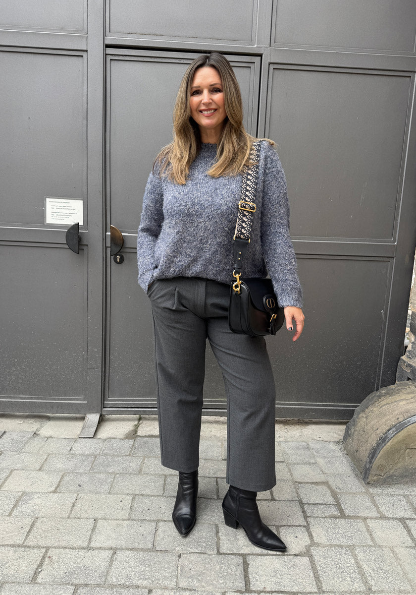 Eine Frau lächelt im Freien auf einem Steinweg vor einer grau getäfelten Tür. Sie trägt einen flauschigen Princess Goes Hollywood-Pullover, eine kurze graue Hose, spitze schwarze Stiefeletten und eine schwarze Crossbody-Tasche mit Kettenriemen.