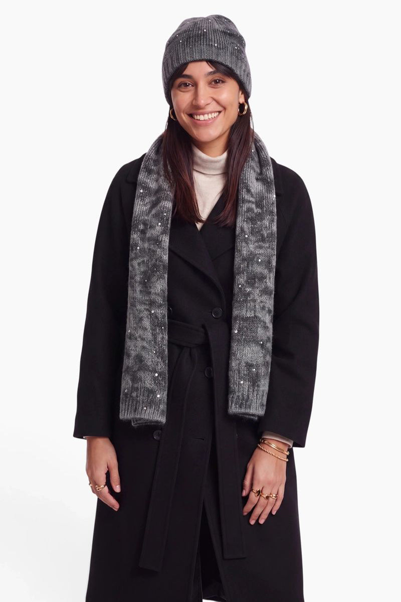Eine lächelnde Frau trägt eine passende graue Mütze und einen Schal aus einer Merino-Kaschmir-Mischung, die mit kleinen silbernen Nieten verziert sind. Sie trägt einen schwarzen Mantel mit Gürtel über einem cremefarbenen Rollkragenpullover und posiert vor einem schlichten weißen Hintergrund.