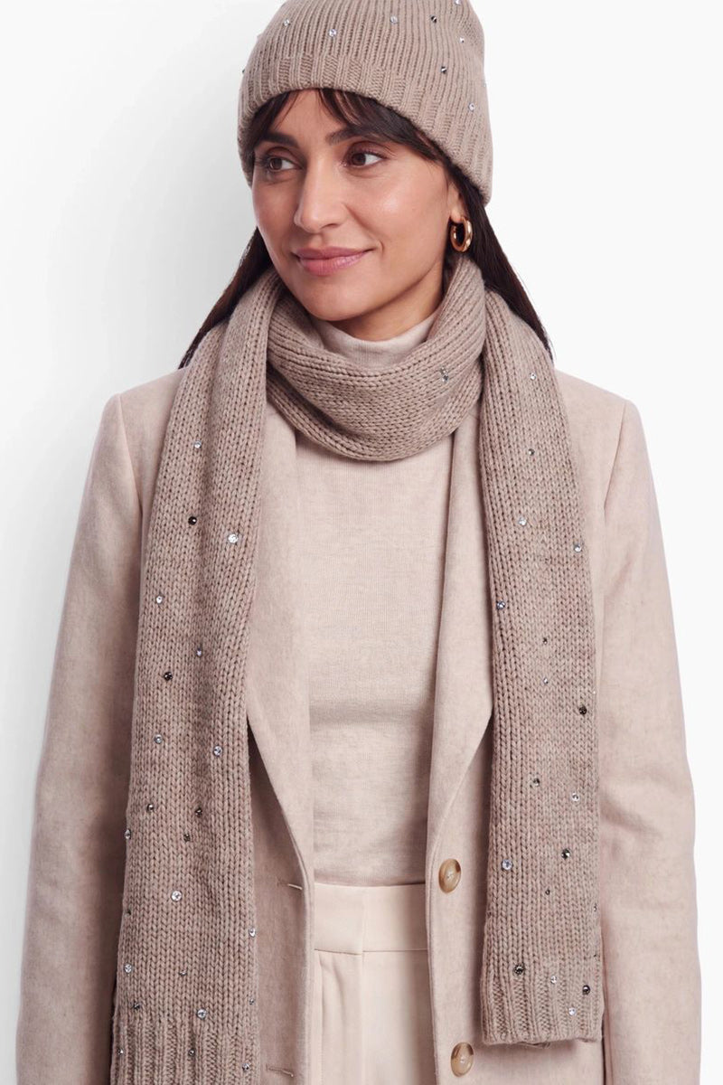Eine Frau trägt eine beigefarbene Mütze aus Merino-Kaschmir und einen dazu passenden langen Schal, die beide mit kleinen funkelnden Kristallen verziert sind. Vor einem schlichten weißen Hintergrund trägt sie einen hellbeigen Rollkragenpullover und einen dazu passenden Wollblazer.
