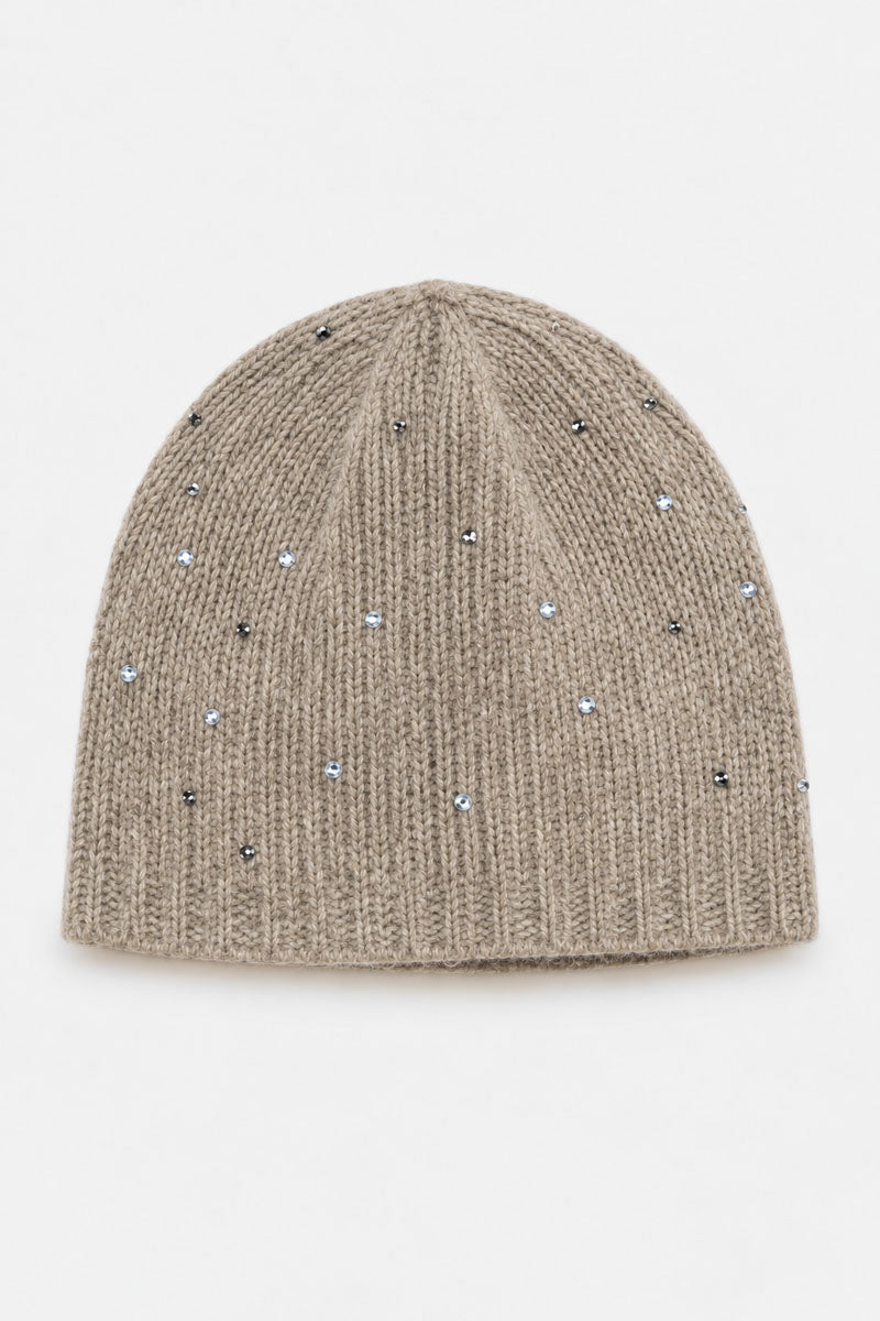 Eine beigefarbene Mütze aus Merino-Kaschmir-Strick, verziert mit vereinzelten, schimmernden silbernen Strasssteinen. Die Mütze hat eine klassische, runde Form und einen gerippten Rand, der sich von einem schlichten weißen Hintergrund abhebt.