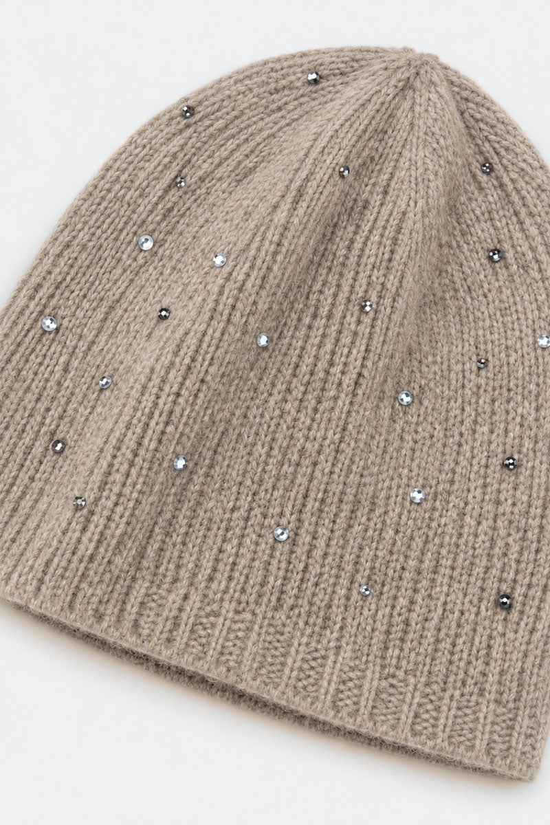 Eine beigefarbene, gerippte Mütze aus Merino-Kaschmir ist mit kleinen, funkelnden silbernen Nieten übersät. Die Mütze hat eine klassisch gerundete Krone und ein strukturiertes Muster auf schlichtem weißem Hintergrund.