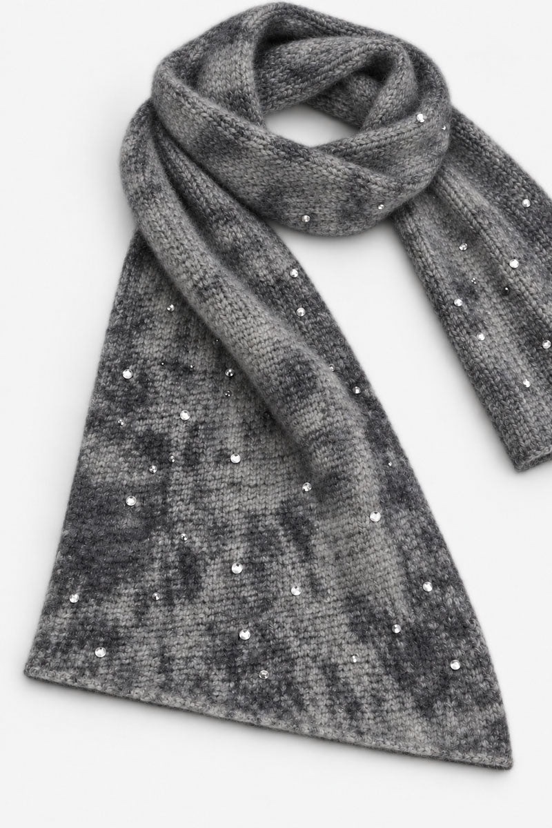 Ein grauer, melierter Strickschal aus einer Merino-Kaschmir-Mischung ist locker zu einer Schleife drapiert. Der flauschige Stoff weist einen dezenten Used-Look auf und ist mit vereinzelten klaren Kristallen verziert, die das Licht auf der Wollmischung einfangen.