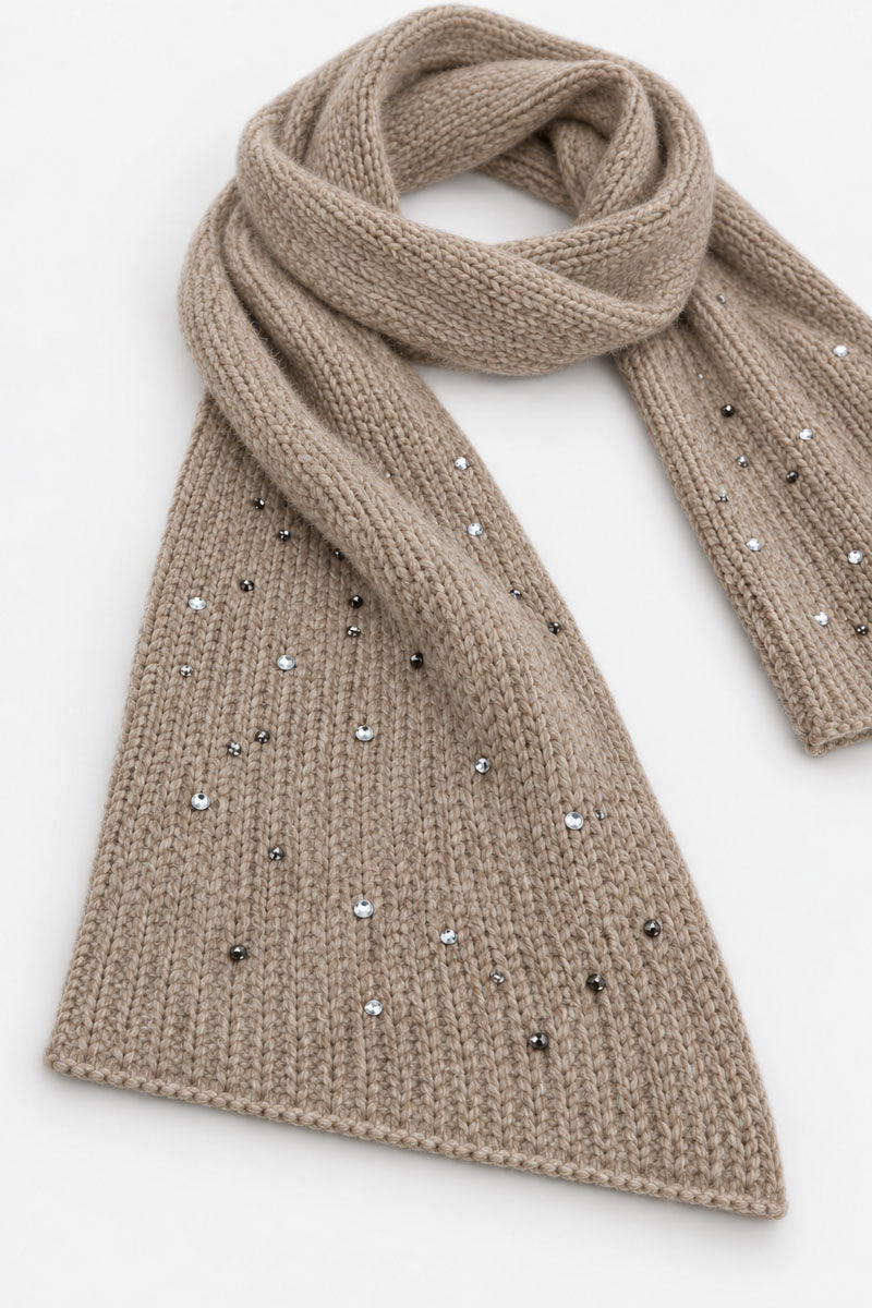 Ein beiger, gerippter Schal aus einer Merino-Kaschmir-Mischung liegt vor einem weißen Hintergrund. Er hat spitz zulaufende Enden und einen individuellen Used-Look mit vereinzelten silbernen und dunkelgrauen Kristallverzierungen.