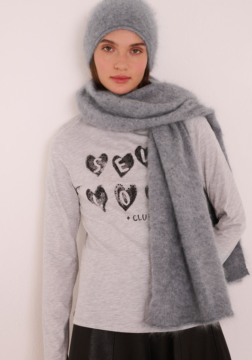 Eine Frau in einer weichen, hellgrauen Princess Goes Hollywood gebürsteten Kaschmir Strickmütze und passendem flauschigen Schal steht vor einer schlichten Wand, trägt ein graues Oberteil mit schwarzen herzförmigen Buchstaben, einen schwarzen Rock und blickt ruhig nach vorne.