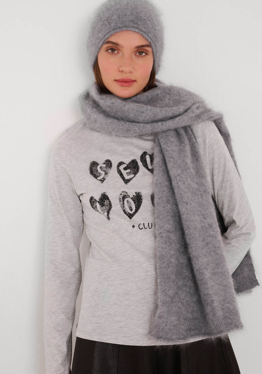 Eine Frau trägt eine weiche graue Princess Goes Hollywood Kaschmir Strickmütze und einen passenden flauschigen Schal mit einem hellgrauen Langarmshirt mit schwarzer Herzgrafik. Sie steht vor einem weißen Hintergrund und schaut in die Kamera.