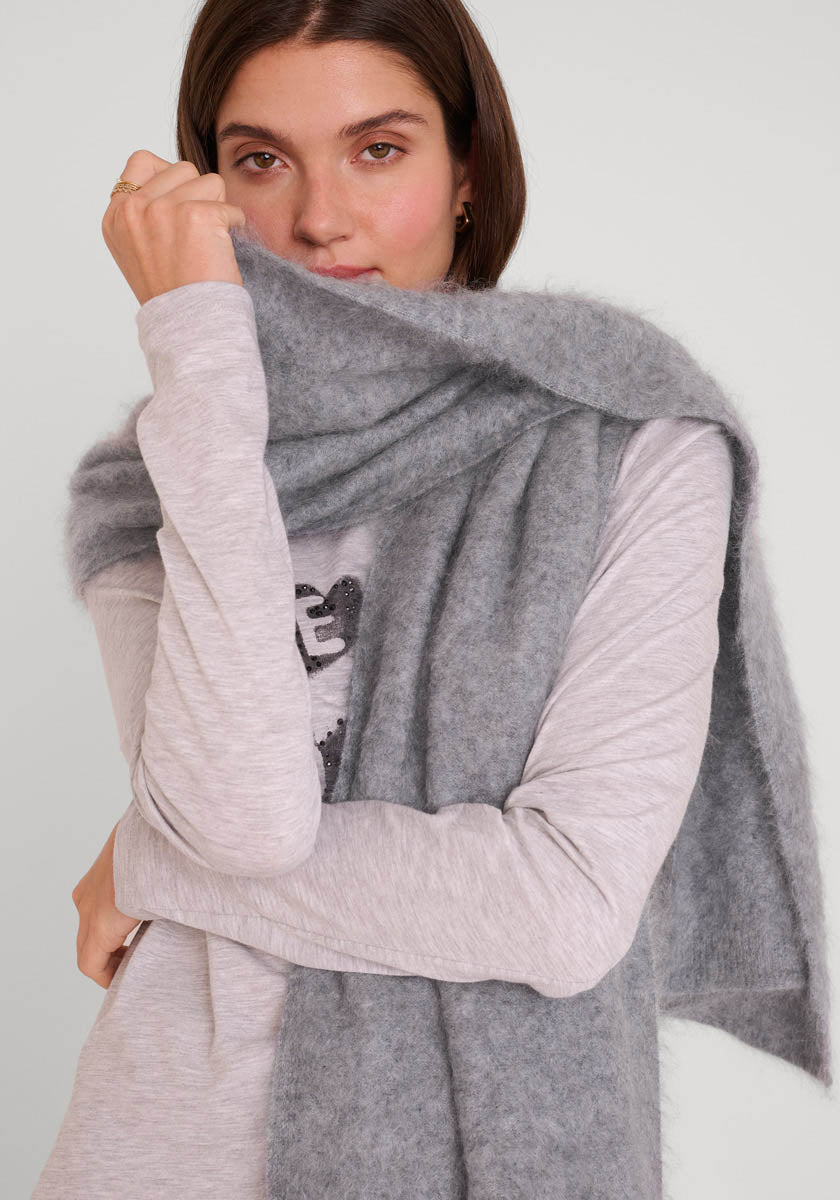 Eine Frau mit glattem braunem Haar trägt ein hellgraues, langärmeliges Oberteil und einen weichen Princess Goes Hollywood gebürsteten Kaschmir Schal. Sie hält sich den kuscheligen Schal vor den Mund und blickt in die Kamera vor einem schlichten Hintergrund.