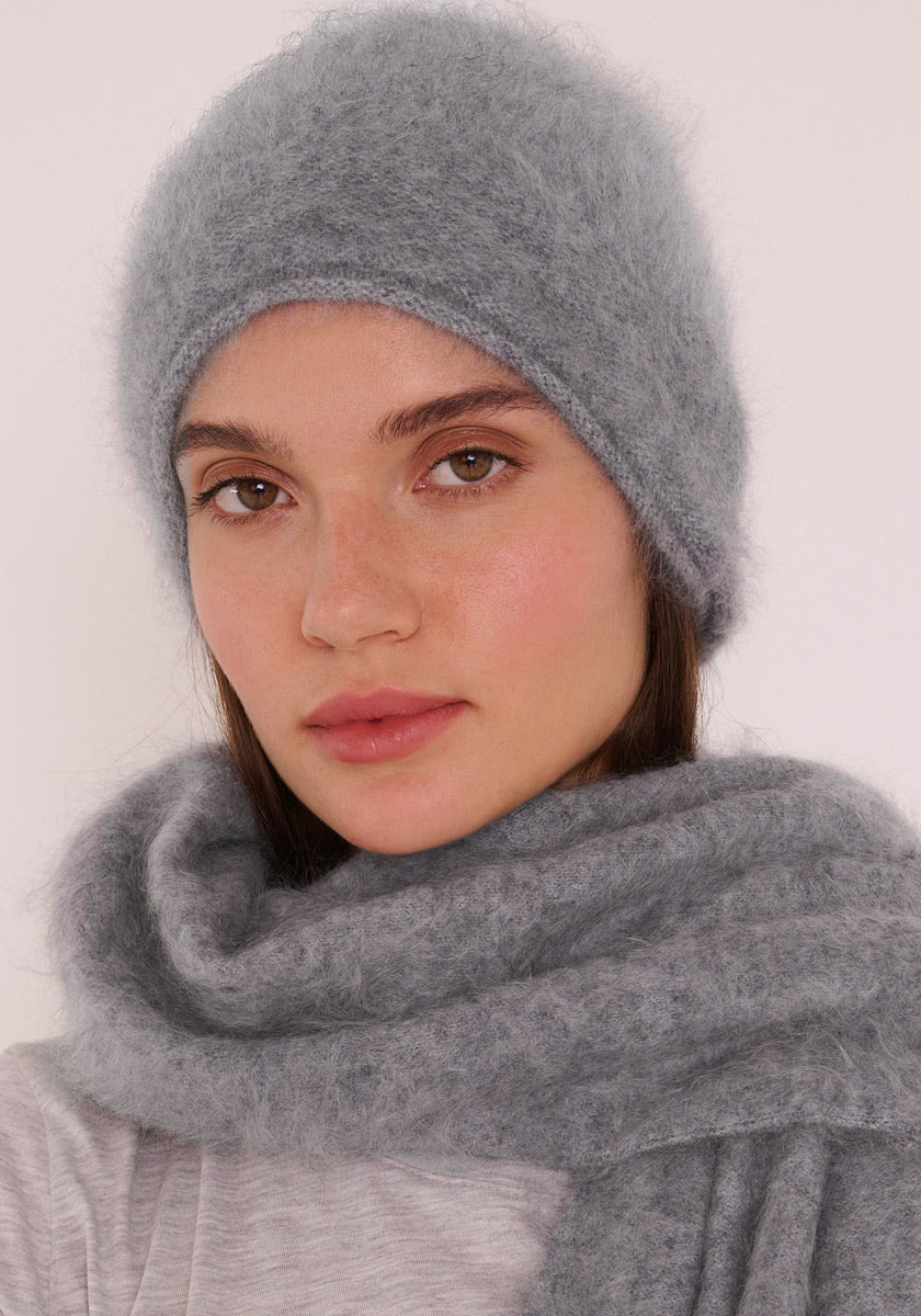 Eine Person trägt eine weiche, flauschige hellgraue Kaschmirmütze und einen passenden Schal von Princess Goes Hollywood. Ihr glattes braunes Haar lugt an den Seiten hervor. Mit heller Haut, natürlichem Make-up und einem neutralen Gesichtsausdruck posiert sie vor einem hellen, unifarbenen Hintergrund.