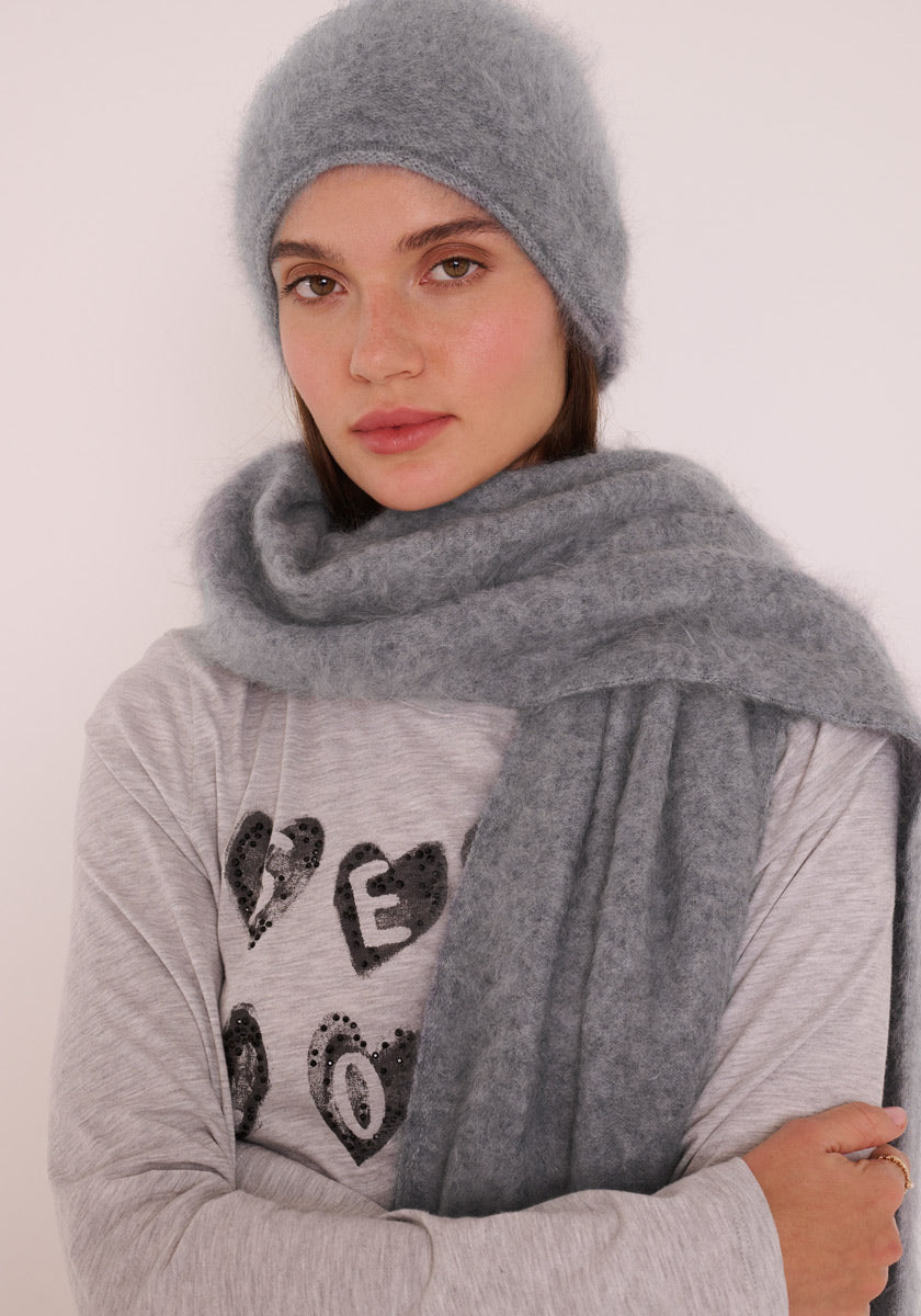 Eine Frau mit heller Haut und glattem braunem Haar trägt eine weiche, gebürstete Kaschmir Strickmütze und einen passenden Schal von Princess Goes Hollywood. Dazu trägt sie ein hellgraues, langärmeliges Shirt mit schwarzem Pailletten-Schriftzug in Herzform.