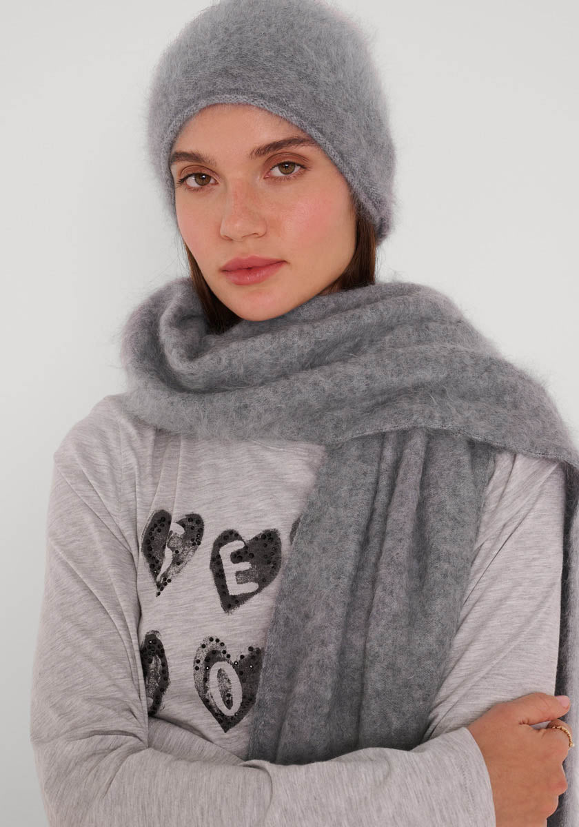 Eine Frau trägt eine weiche graue Kaschmir-Strickmütze und einen passenden Schal von Princess Goes Hollywood vor einem hellen Hintergrund. Ihr langes braunes Haar umrahmt ihr Gesicht. Dazu trägt sie ein graues Shirt mit schwarzen Herzmotiven und Schriftzügen.