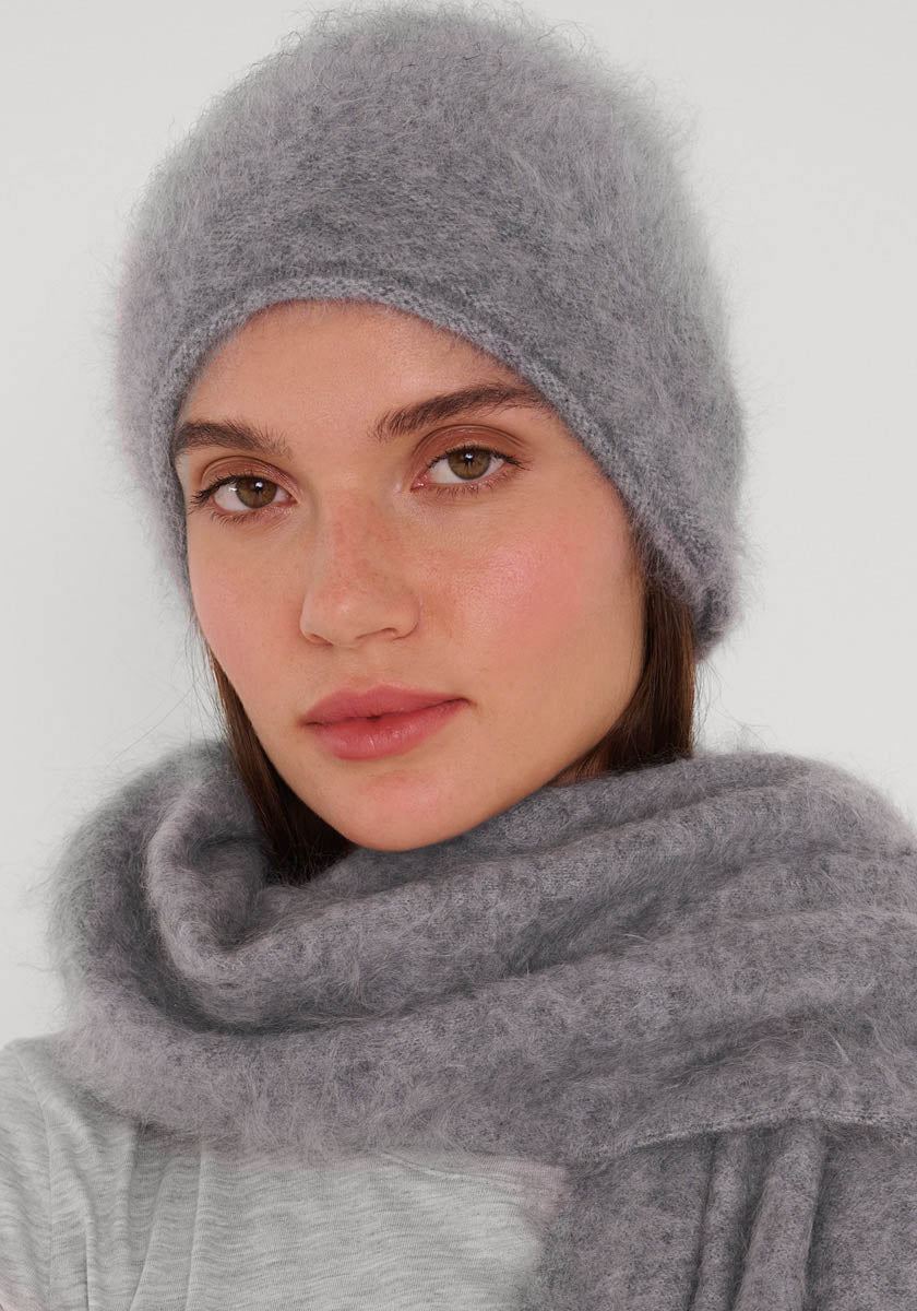 Eine Frau mit heller Haut und braunen Augen modelliert eine gemütliche Princess Goes Hollywood Kaschmir Strickmütze und einen passenden Schal. Sie wird von den Schultern aufwärts gezeigt, hat einen neutralen Ausdruck und steht vor einem schlichten, hellen Hintergrund.