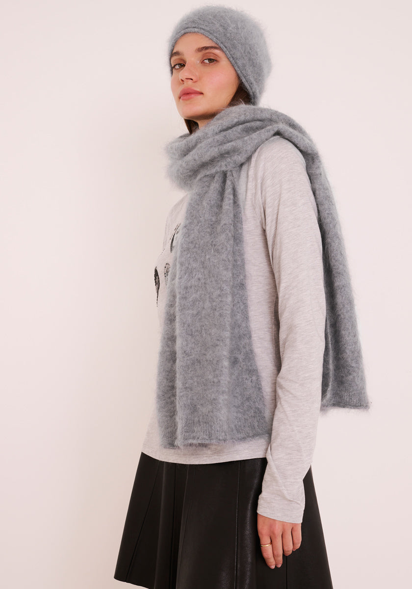 Eine Frau steht vor einem hellen Hintergrund und trägt eine graue Kaschmir-Mütze von Princess Goes Hollywood mit passendem Schal, ein hellgraues langärmeliges Hemd und einen schwarzen Rock aus Kunstleder. Sie schaut mit neutralem Gesichtsausdruck in die Kamera, dunkles Haar unter der Mütze.