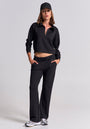 A woman in a black cropped zip-up sweatshirt, elastic-waist sweatpants, black cap, and silver sneakers from Princess Goes Hollywood stands confidently against a plain background, left hand in pocket and gazing forward relaxedly.