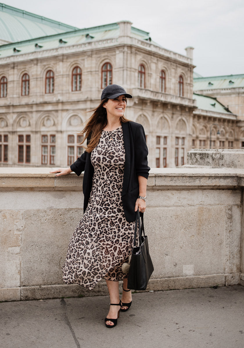 A woman wearing a leopard print dress and black blazer from Princess Goes Hollywood with black Kappe smiles by a stone railing. She completes her outfit with black sandals, a tote bag, and poses before an ornate building with arched windows.