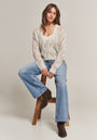 A woman with wavy brown hair sits on a wooden stool, wearing a cream-colored Cardigan with Lurexfäden by Princess Goes Hollywood over a white top, light blue wide-leg jeans, and dark brown ankle boots. She looks relaxed against a plain light background.