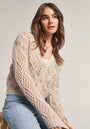 A young woman with wavy brown hair sits on a stool, wearing a beige diamond-patterned Merino-Kaschmir-Mix Ajourstrick cardigan by Princess Goes Hollywood and light blue jeans. She looks slightly upward, her expression soft against a plain background.
