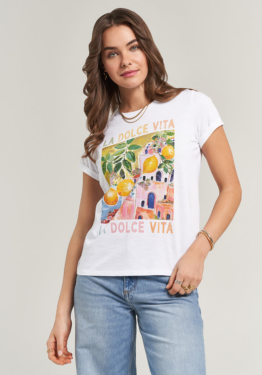 A woman with wavy brown hair models the Princess Goes Hollywood T-Shirt La Dolce Vita mit Zitronenprint und pastellfarbenen Gebäuden, kombiniert mit hellblauen Jeans und goldfarbenem Schmuck vor hellem Hintergrund.