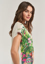 A woman with long brown hair stands in profile, smiling gently as she wears the cream-colored T-Shirt Garden by Princess Goes Hollywood, featuring a vibrant garden print and classic round neckline against a light gray background.