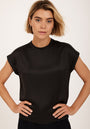 A woman with short blonde hair stands facing forward, hands on hips, wearing a plain black Kurzarmbluse by Princess Goes Hollywood and small hoop earrings. The light background emphasizes her confident expression and modern style.