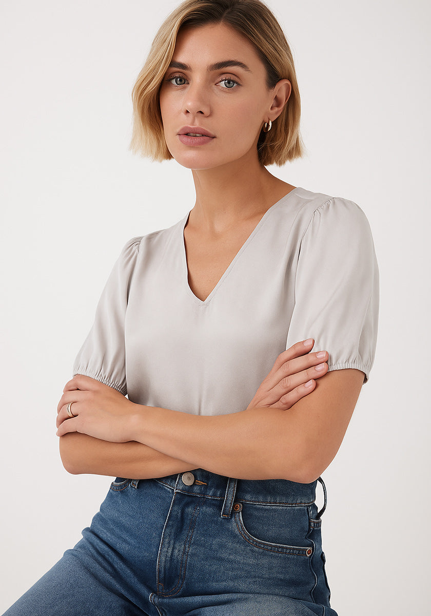 A woman with short blonde hair and light skin wears a pale gray Princess Goes Hollywood Seidenbluse mit V-Ausschnitt und kurzen Ballonärmeln, paired with blue high-waisted jeans, standing calmly with crossed arms against a plain light background.