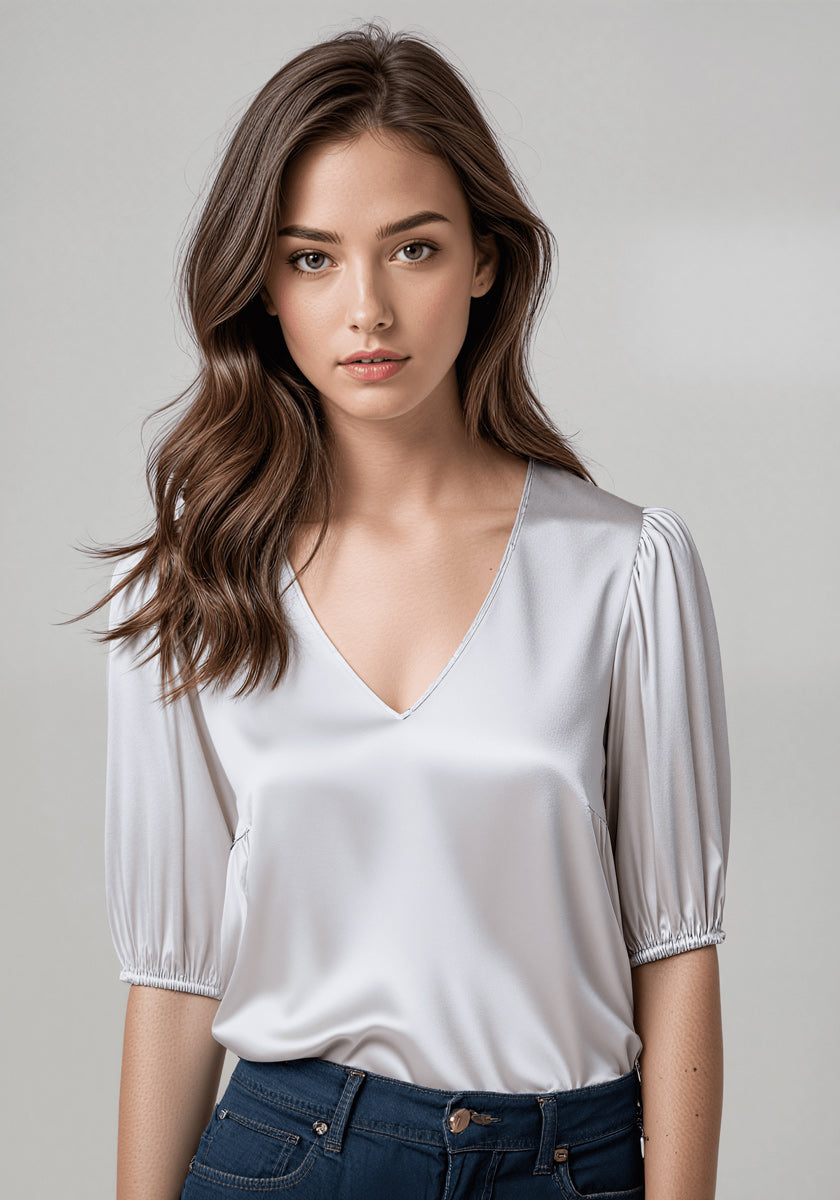 A young woman with long, wavy brown hair models a Princess Goes Hollywood weiße Seidenbluse mit V-Ausschnitt und kurzen Ballonärmel und dunkle blaue Jeans. Sie steht vor grauem Hintergrund und blickt neutral in die Kamera.