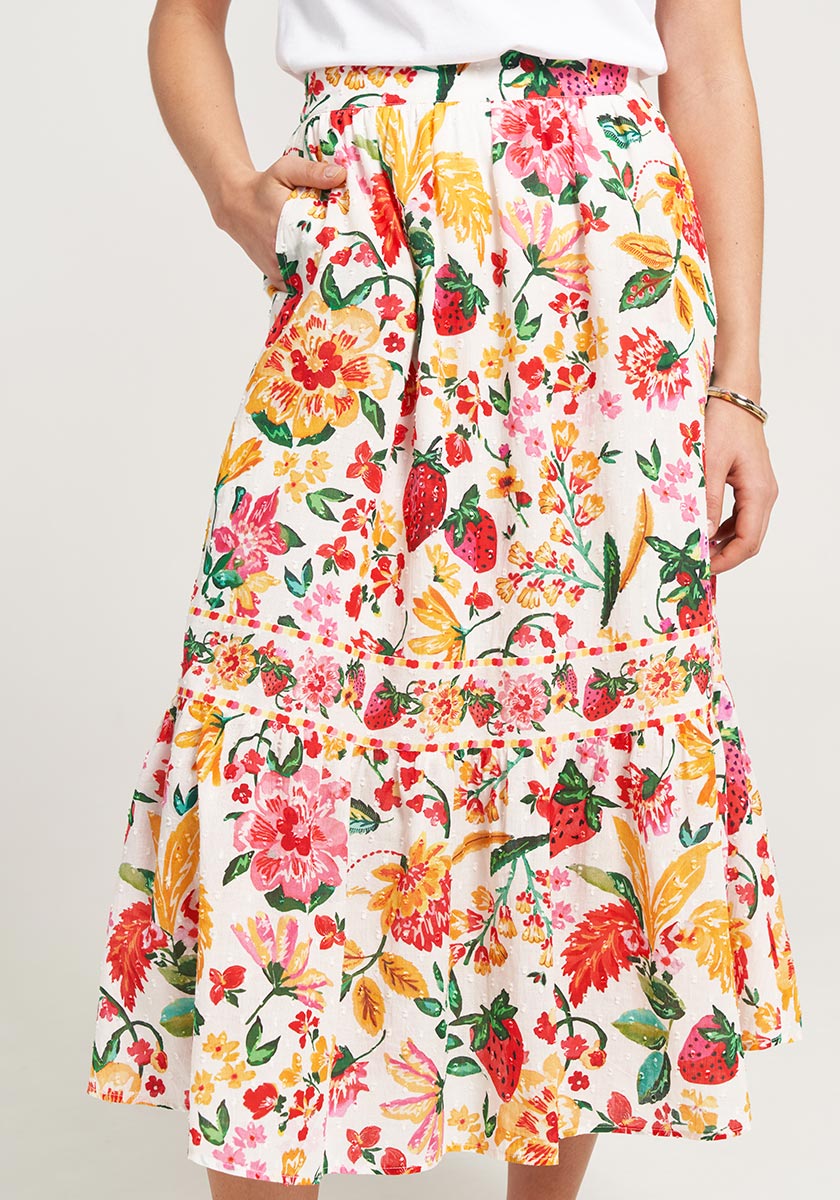 A woman wears a Princess Goes Hollywood cotton midi skirt with a vibrant floral allover print in red, yellow, pink, and green. The mid-length skirt has pleats, ruffled hem, and pockets. She pairs it with a tucked-in white top and a slim bracelet.