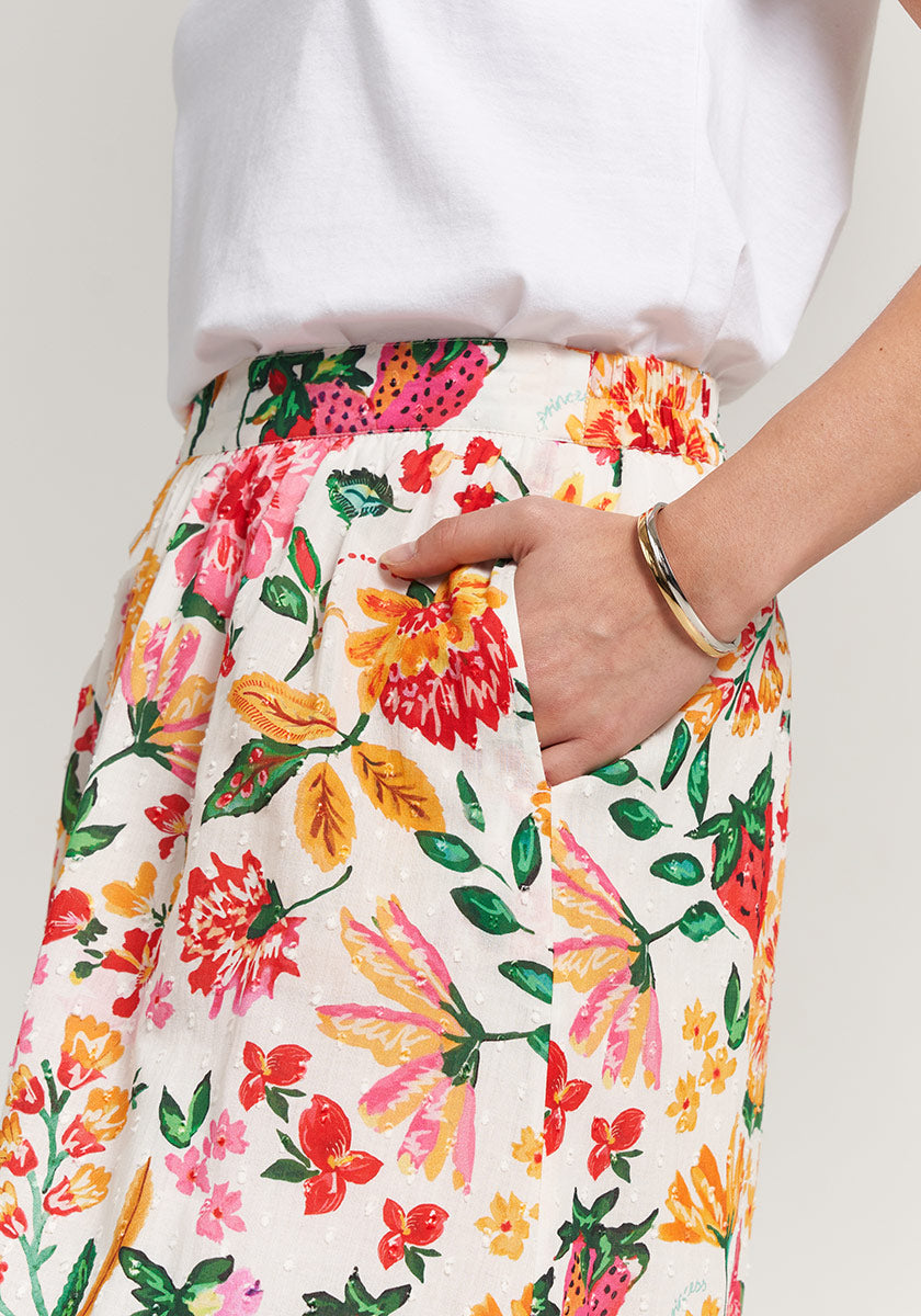 A person wears a white short-sleeve top and Princess Goes Hollywood high-waisted floral pants with vibrant red, pink, yellow, and green patterns. Their right hand is in the pocket, complemented by a silver bracelet.